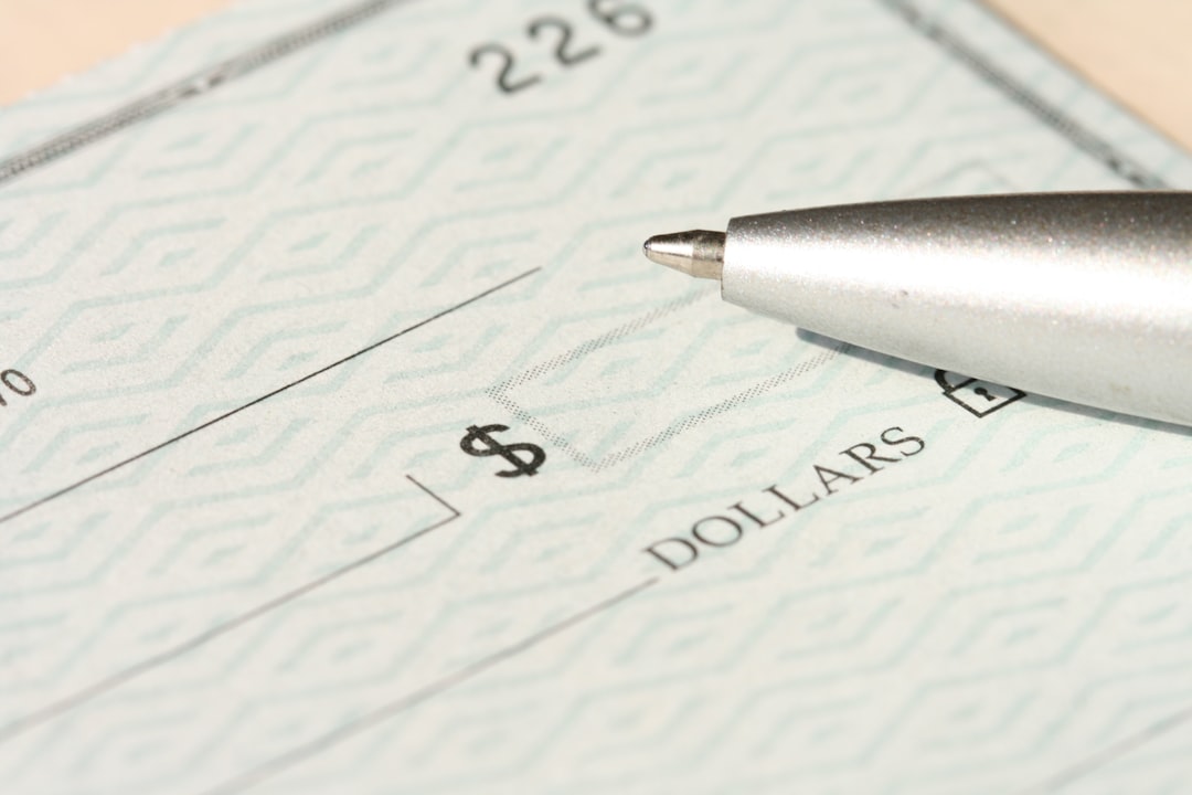 A close-up, high-resolution image of a blank check with a security pattern, showing the dollar sign box and part of the “DOLLARS” line. A metallic ballpoint pen rests near the writing area.